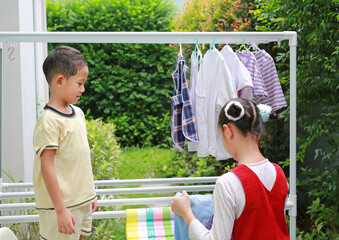 Portrait of Asian girl and boy helping to do dry the clothes at garden near house. Sister and little brother are help to housework. Kids hanging the laundry on a clothesline rail. - 761951666