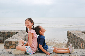 Portrait of cute Asian young girl child and little boy kid sitting together at seacoast. Sister and brother sit back against each other on cement wall. - 761951801