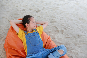 Portrait of Asian young girl in dungarees jean looking beside and relax on orange sofa bed beach on sand at summer holiday. - 761953231