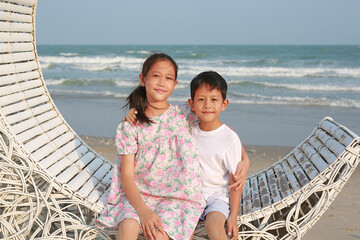 Portrait of happy Asian boy and girl sitting and hugging each other on white bamboo wooden sofa bed and looking camera on the beach at summer holiday. - 761953411