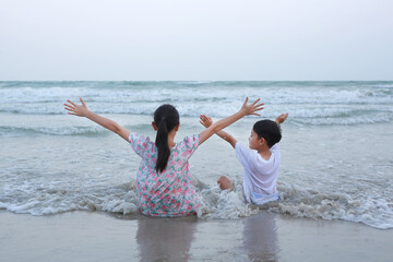 Back view of Asian young girl child and little brother sitting and raised hands up on tropical sand beach at sunset. Adorable sister and brother having fun in summer holiday. - 761953442