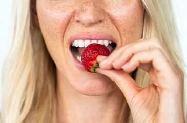 Blonde woman with freckles smiling biting on a strawberry
