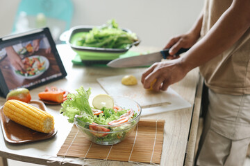 Corn and salad in a bowl with man cutting lemon while watching online cooking course via laptop on the background