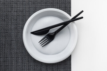 Plastic, disposable white plate, black knife and fork on a black and white background. Top view, studio shot.
