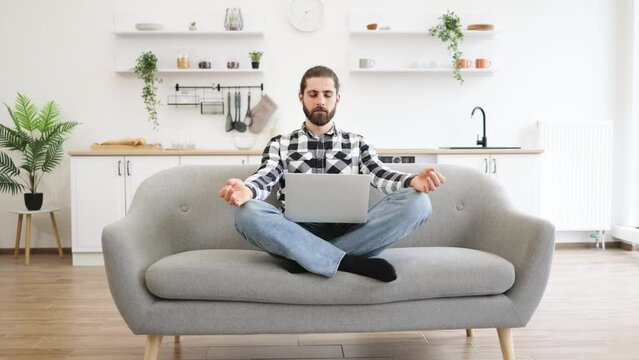 Peaceful Caucasian businessman in casual attire sitting on couch with wireless laptop on knees and meditating. Bearded young guy with fingers in mudra gesture relieve stress and regaining energy.