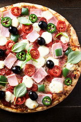 Pizza with ham, mozzarella cheese, cherry tomatoes, green and jalapeno pepper, black olives and fresh basil. Dark background. Top view. Close up.	