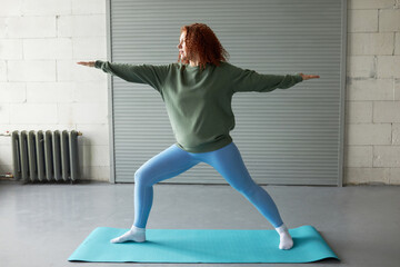 Side view of redhead female in leggings and sport sweatshirt standing on mat with outstretched...