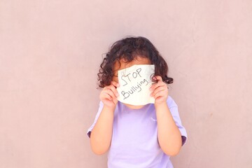 A toddler girl stands holding up a piece of paper with the words stop bullying written on it.