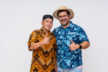 An Indonesian man in batik and kopiah hat smiling with a tourist in a Hawaiian shirt, both giving thumbs up, isolated on white.