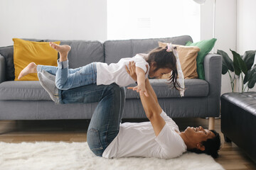 Joyful young father lying on carpet floor, lifting excited happy little daughter at home. Father...