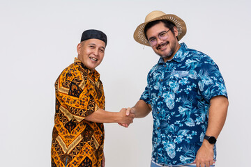 A smiling Indonesian man in traditional batik attire and kopiah hat warmly greets a tourist in a vibrant hawaiian shirt, handshake gesture captured on white background.