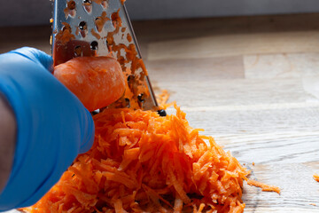 Person, wearing rubber gloves, grating fresh raw carrot with a metal grater in a kitchen