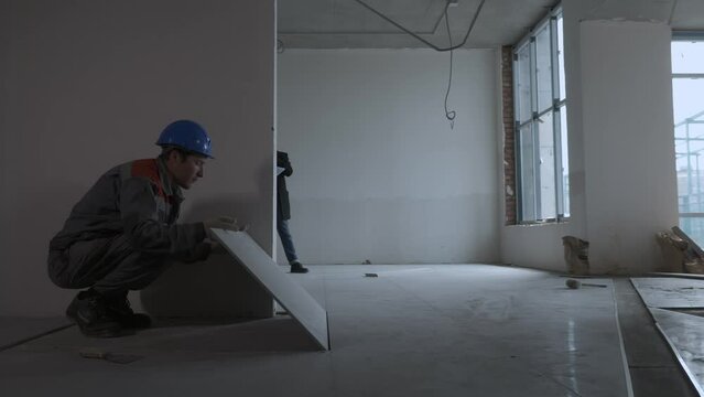 Worker laying tiles. In the next room, engineers are discussing the construction plan
