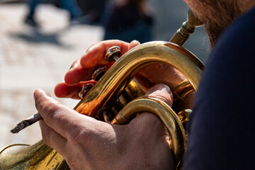 Man's hand playing an old, worn trumpet in an outdoor park while holding a lit cigarette