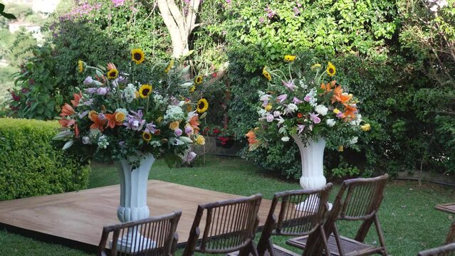 A beautiful garden with two vases of flowers with bamboo chairs around them, set for a wedding celebration