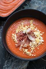 Spanish traditional salmorejo served with prosciutto and grated egg in a black bowl, vertical shot, middle close-up
