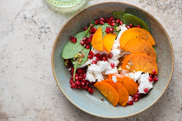 Persimmon with goat cheese, spinach, pomegranate and pistachios in a rustic plate, horizontal shot on a beige granite background, high angle view