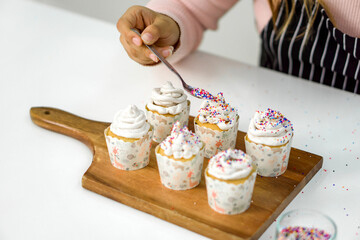 Closeup woman hand holding spoon with multicolor frosting, decorating cupcake on a wooden board, adding a colorful touch to the cupcakes' presentation.