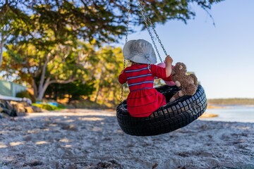 young girl toddler in red dress on a tyre swing on a sandy beach with soft toys