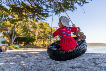 young girl toddler in red dress on a tyre swing on a sandy beach with soft toys