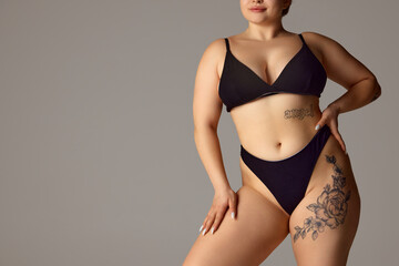 Cropped portrait of female plus-size model posing in dark bikini, underwear against grey studio...