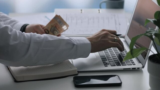 A young man writes financial expenses and income in a notebook. Control of banking operations. There is a laptop, a notebook, a folder with documents, a smartphone and cash on the table.