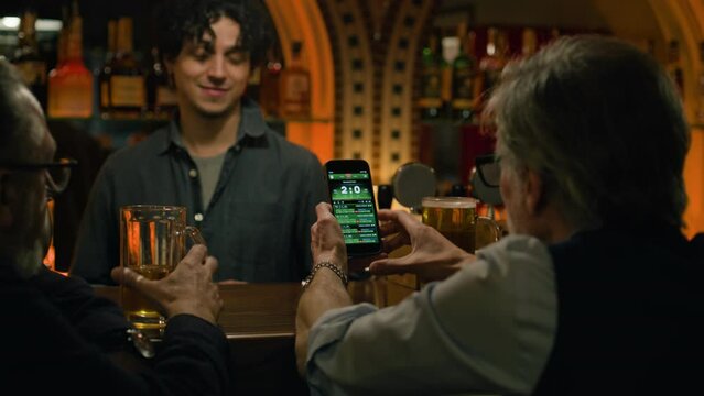 Mature sports fans check game score and bookmaker ratings in app using mobile phone, talk with bartender. Male friends bet on football match online sitting at bar counter in pub. Gambling concept.