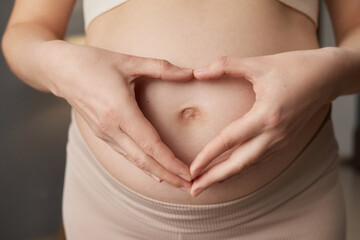 Loving unrecognizable pregnant woman with bare belly standing in home interior making heart shape with hands on her tummy expressing gentle wearing beige clothing