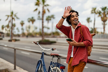 A happy trendy guy waving to a friend and holding cellphone on a street. - obrazy, fototapety, plakaty