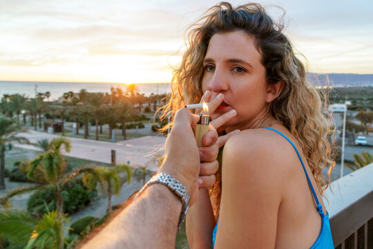 Young woman with long wavy hair in a blue swimsuit holds a cigar in her hands and looks at the sunset