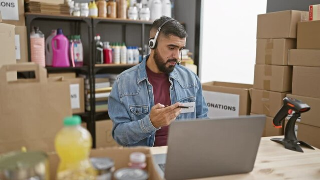 Bearded man with headset in warehouse filled with donation boxes and household items browsing laptop.