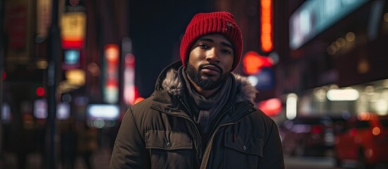 A man with a red hat and black jacket stands on a city street at night, surrounded by darkness. His facial hair is visible under the dim streetlights - obrazy, fototapety, plakaty