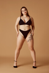Fototapeta premium Young confident, beautiful woman posing in black bikini and stands on heels against sandy color studio background. Model plus-size. Concept of natural beauty, femininity, body positivity, dieting.