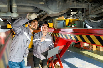Two mechanic in gray uniform maintenance or inspection work on a vehicle that is raised on a lift while holding laptop computer. Diagnostic, referencing repair manual, or updating service record.