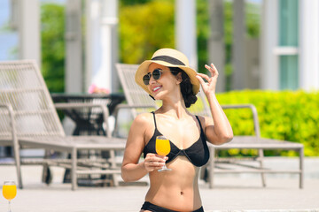 Happy smiling woman in bikini with straw hat relaxing with orange juice at poolside. beautiful female relaxing in summer time. holiday vacation concept