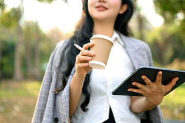 Satisfied young businesswoman drinking coffee and using digital tablet in the park