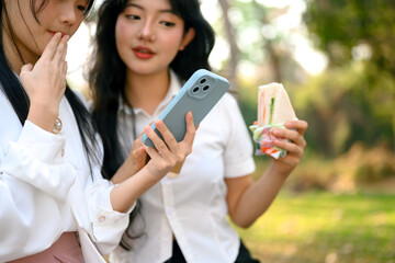 Happy female friends eating sandwich and checking news feed or messaging mobile phone at outdoor