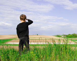 7 year old boy in black tracksuit looks in distance on background of beautiful green agricultural field.Wheat field. Agriculture.