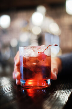 A close-up image capturing a moment of someone holding a glass of cherry cocktail, garnished with a cherry, at a well-lit bar