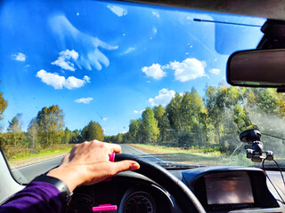 view from a car windshield of natural landscape with road, green trees and blue sky in summer or spring time. Hand of woman on the steering wheel. Female traveler driving alone on trip or journey