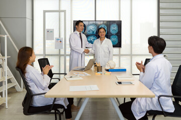Two doctors shaking hands happily in a meeting with colleagues clapping.