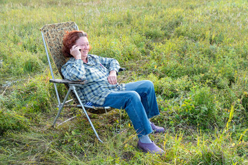 A woman in a folding chair is talking on the phone in a green meadow