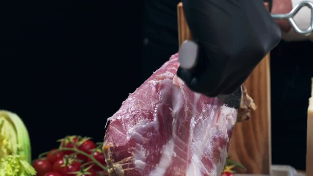 Preparation of Italian antipasto for catering service, chef in black gloves slicing cured prosciutto for degustation, person cutting off fat parts of cured ham before meat slicing, traditional