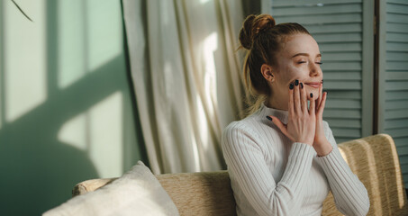 Fototapeta na wymiar Image of young woman applying a cosmetic cream mask on face, sitting on sofa enjoying her leisure time, relaxing alone. Best skin moisturizer. Beauty skin