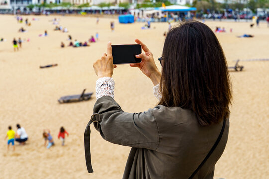 A woman is capturing a moment on her smartphone, taking a photograph of a lively beach scene where people sunbathe and play in the sand.
