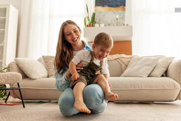 mother is holding her little son in her arms and having fun at home, 2 year old boy is playing with...