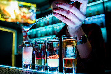 bartender skillfully pouring a layered shot at a bar. Various filled shot glasses adorn the bar,...