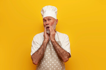 shocked old bald grandfather chef in hat and apron looks surprised on yellow isolated background