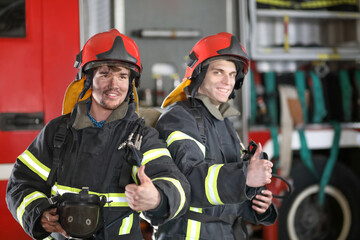 Two firefighters in protective suits with half mask in hand against fire engine, gesture thumbs up