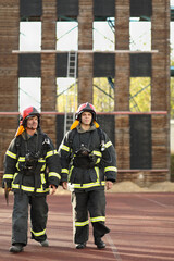 Two firefighters in equipment and red helmets go on training test area against fire tower